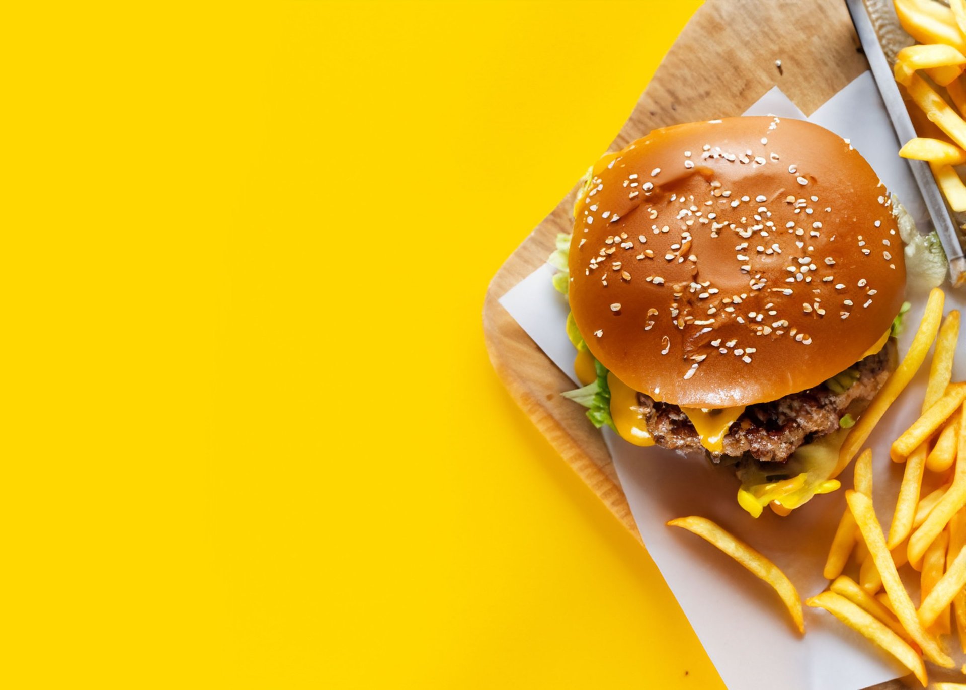 Burger with sesame seed bun and crispy french fries on a white plate against a bright yellow background