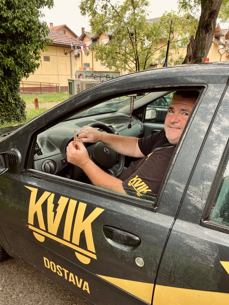 Man sitting in drivers seat of black and yellow delivery van with KVIK Dostava branding, smiling at camera