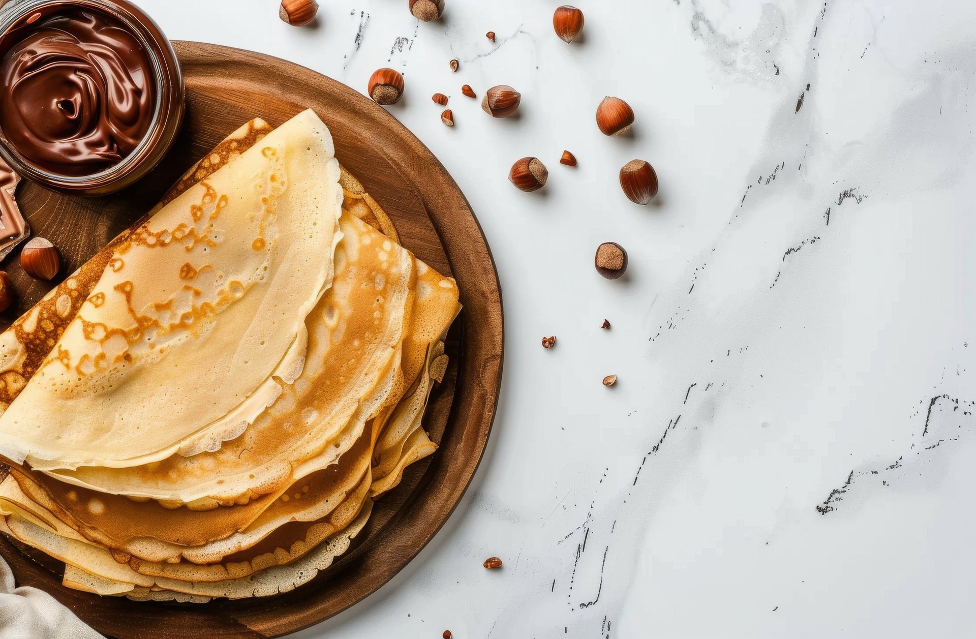 Stack of crepes on a wooden plate with chocolate spread and scattered hazelnuts on white marble surface