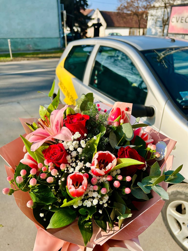 Bouquet of red roses, pink lilies, and white babys breath wrapped in pink paper, held outdoors near a yellow and white car