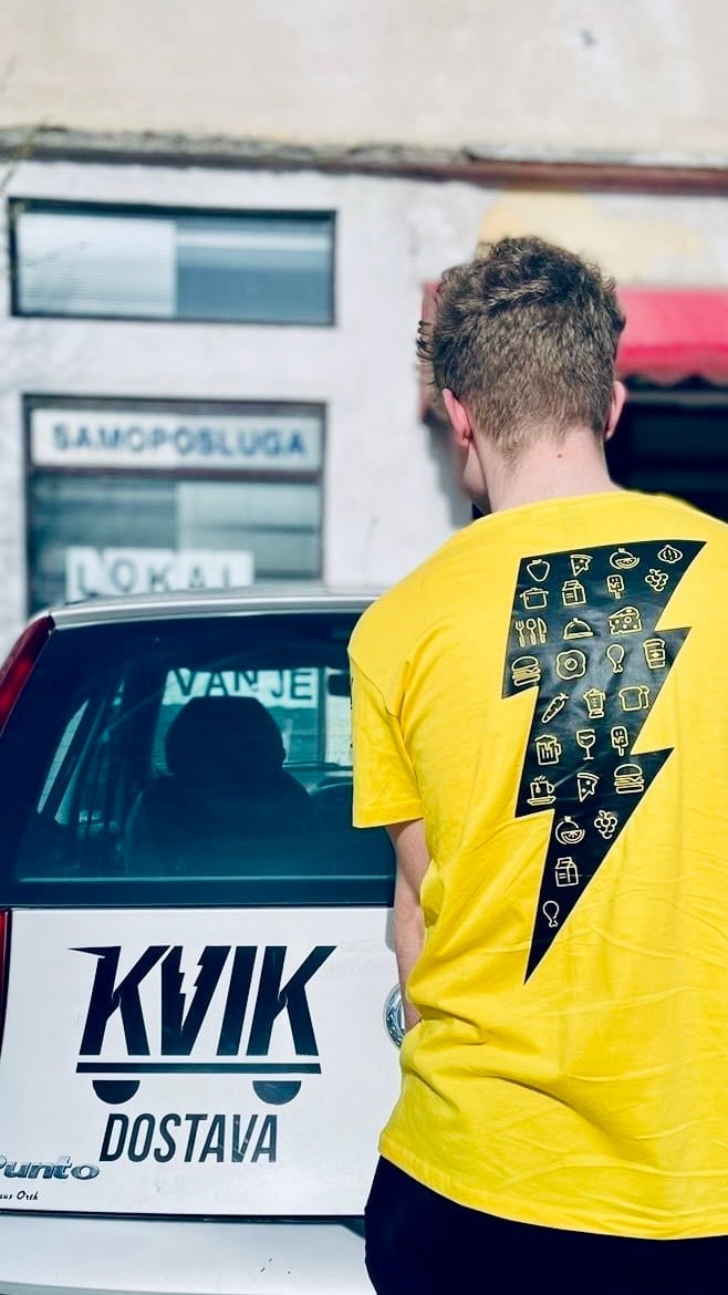 Person wearing yellow t-shirt with graphic design standing next to delivery van with KVIK DOSTAVA branding and storefront window